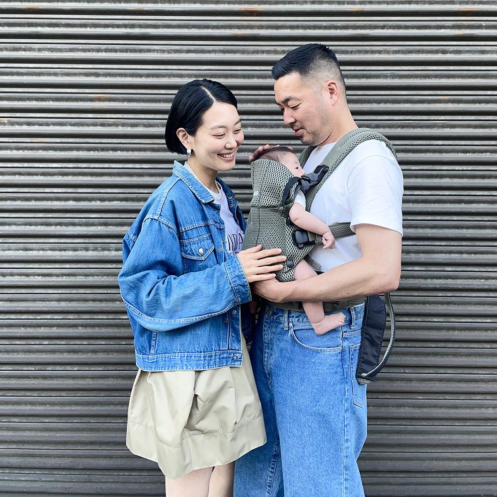 A smiling couple stands in front of a metal shutter, with the man holding a baby in a carrier. The woman gently touches the baby's arm.