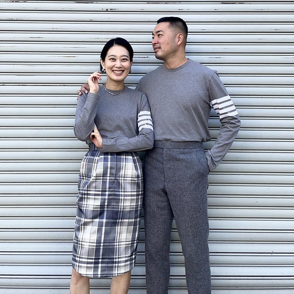 A smiling couple in matching gray tops stands in front of a corrugated metal wall. The woman wears a plaid skirt, and the man wears gray pants.
