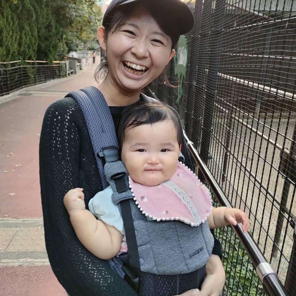 A smiling woman wearing a cap holds a baby in a carrier on a sidewalk, both looking happy. The baby wears a pink bib.
