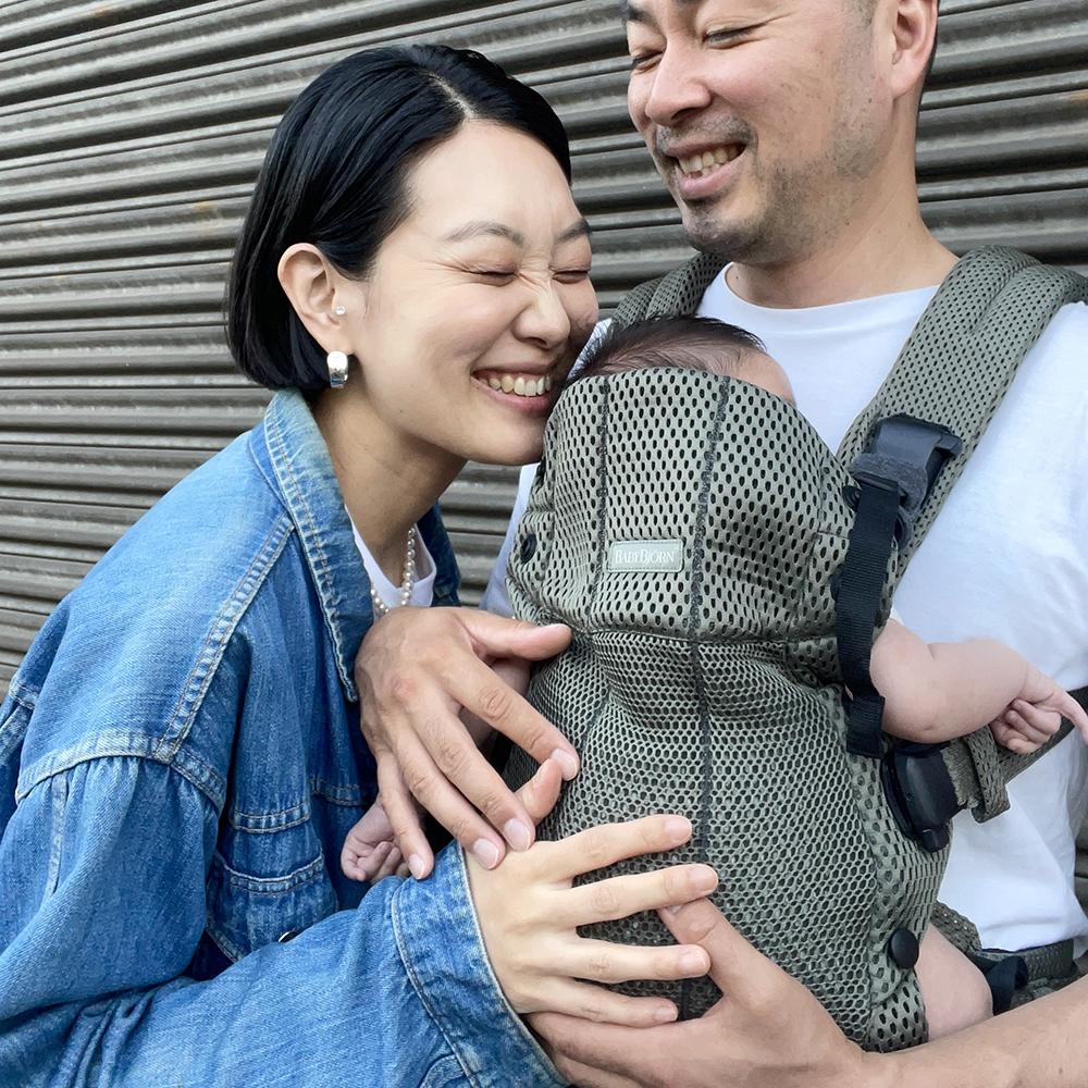 Smiling couple with a baby in a carrier. The woman wears a denim jacket, and the man holds the baby, standing in front of a metal shutter.