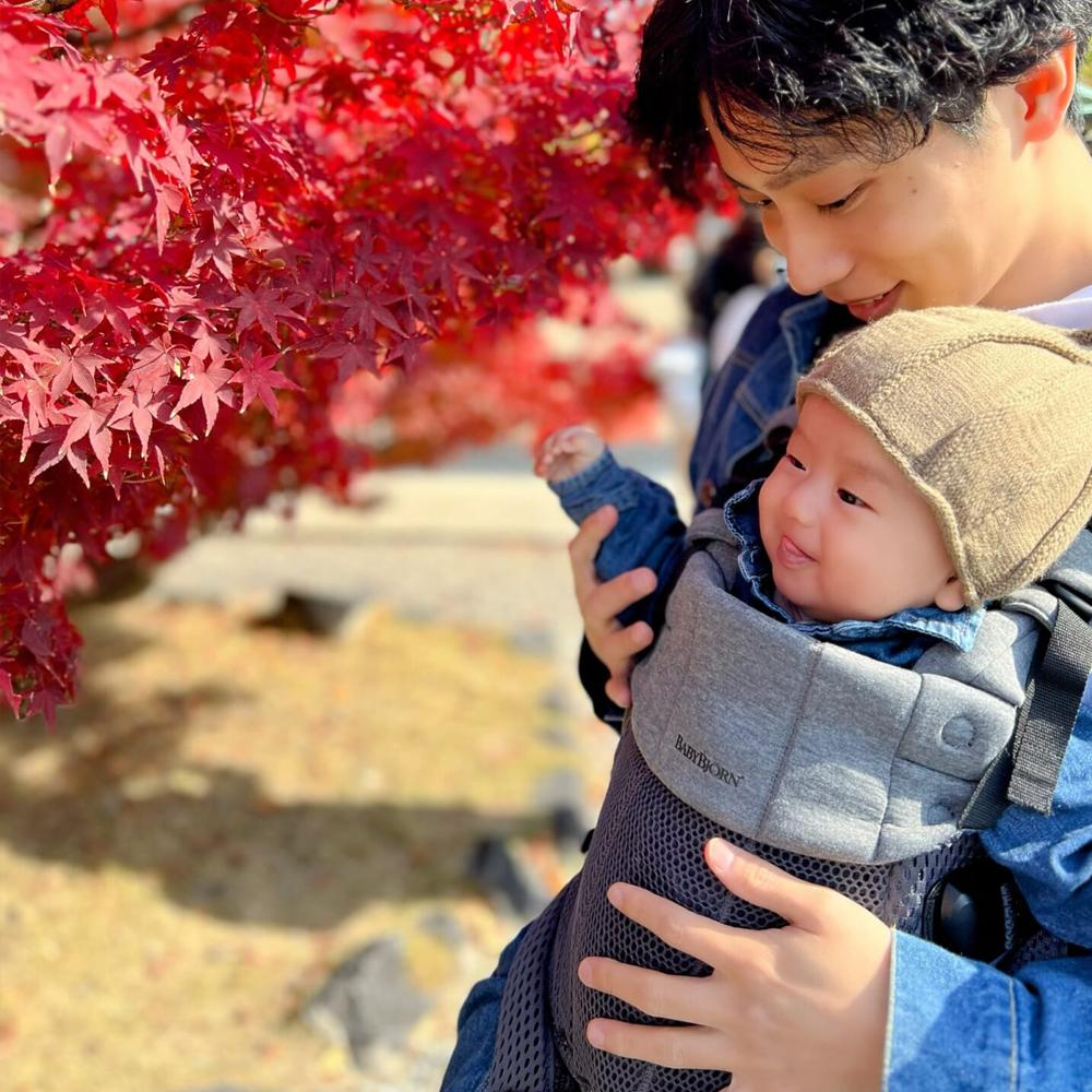 A person holds a smiling baby in a carrier, standing near a vibrant red maple tree. Both are wearing blue clothing.