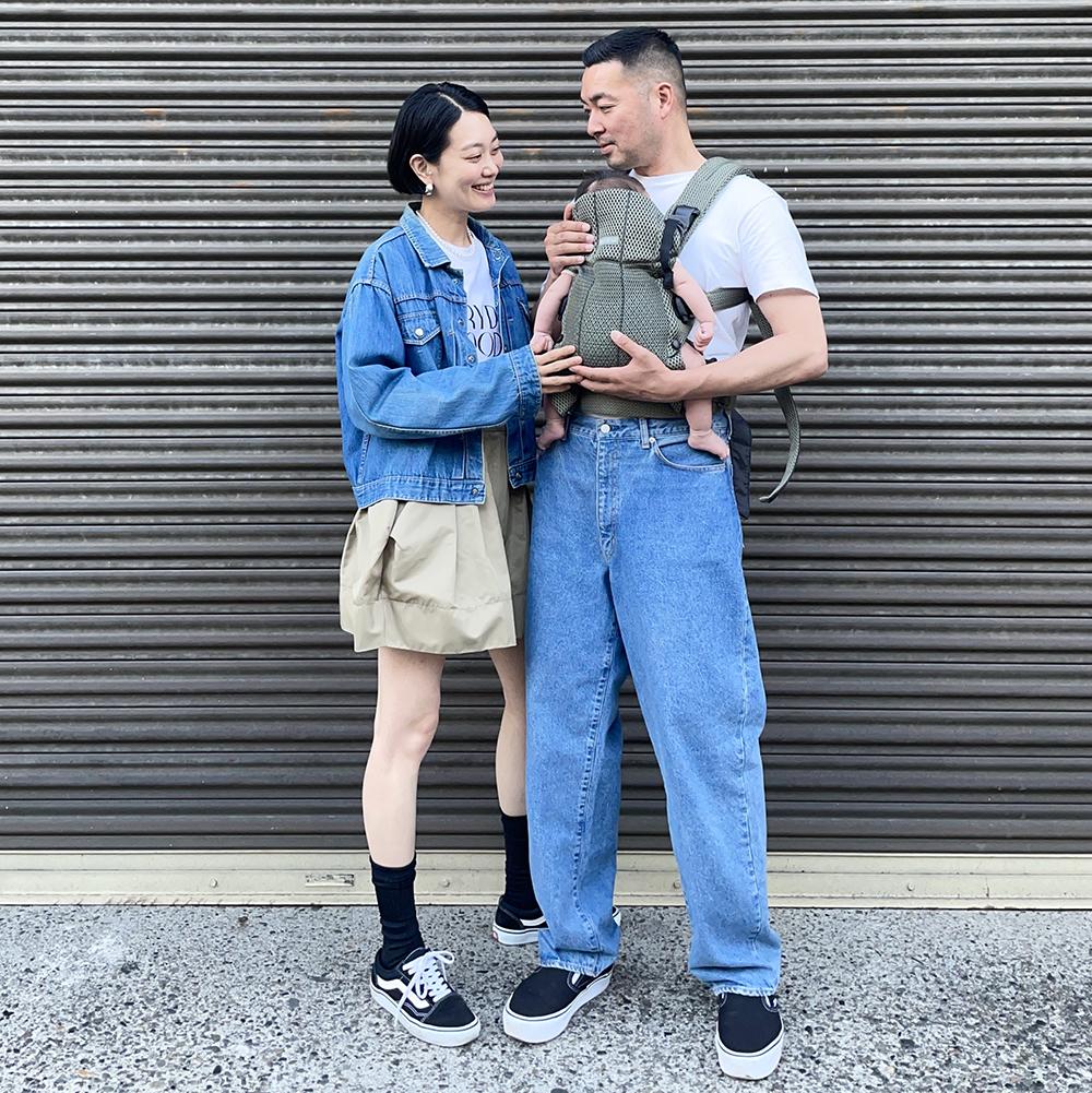 A couple stands smiling at each other, with the man holding a baby in a carrier. They're in casual attire in front of a metal shutter.