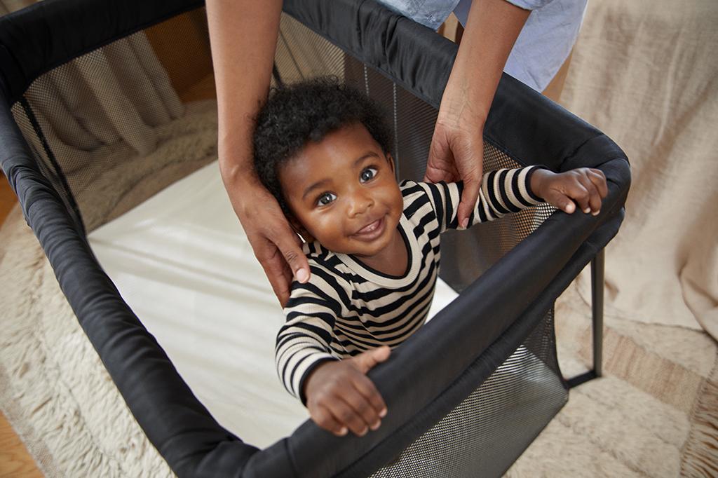 A smiling baby in a striped outfit stands in a playpen, holding the edge, with an adult's hands gently supporting them.