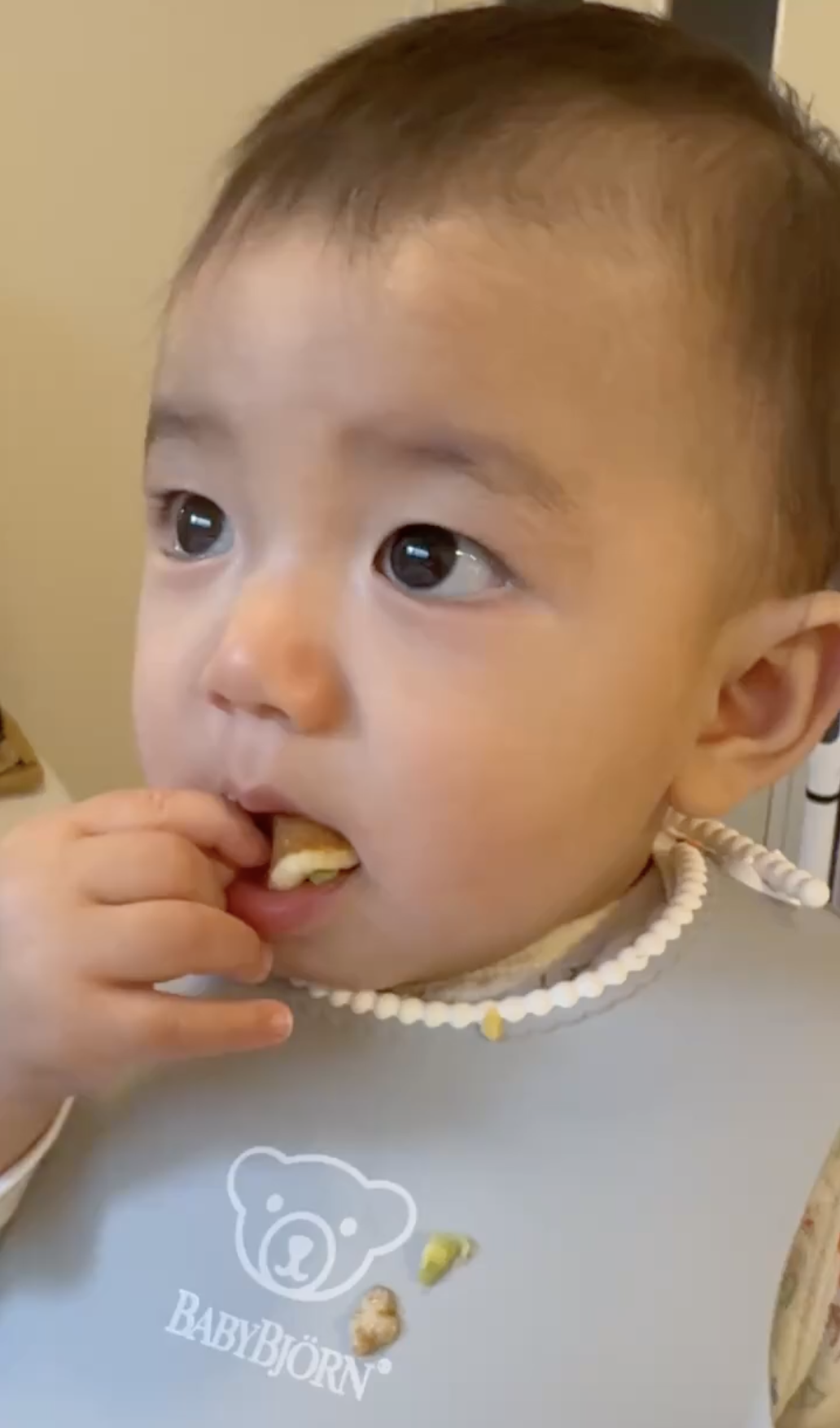 A baby wearing a bib is eating, with food on their fingers and a piece in their mouth, looking slightly to the side.