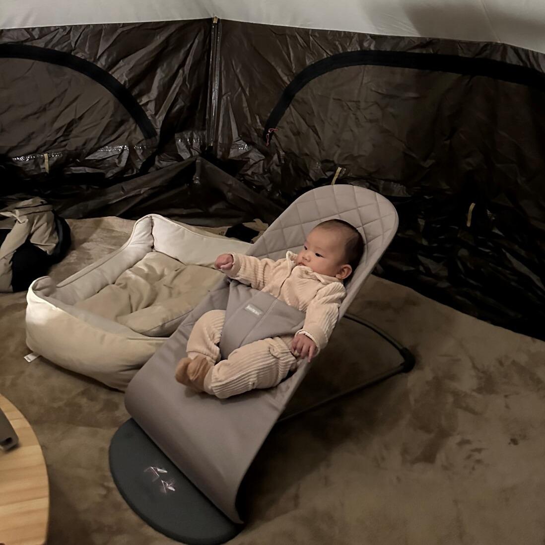 Baby in a cozy chair inside a tent, dressed in a light-colored outfit, surrounded by camping gear on a soft carpet.
