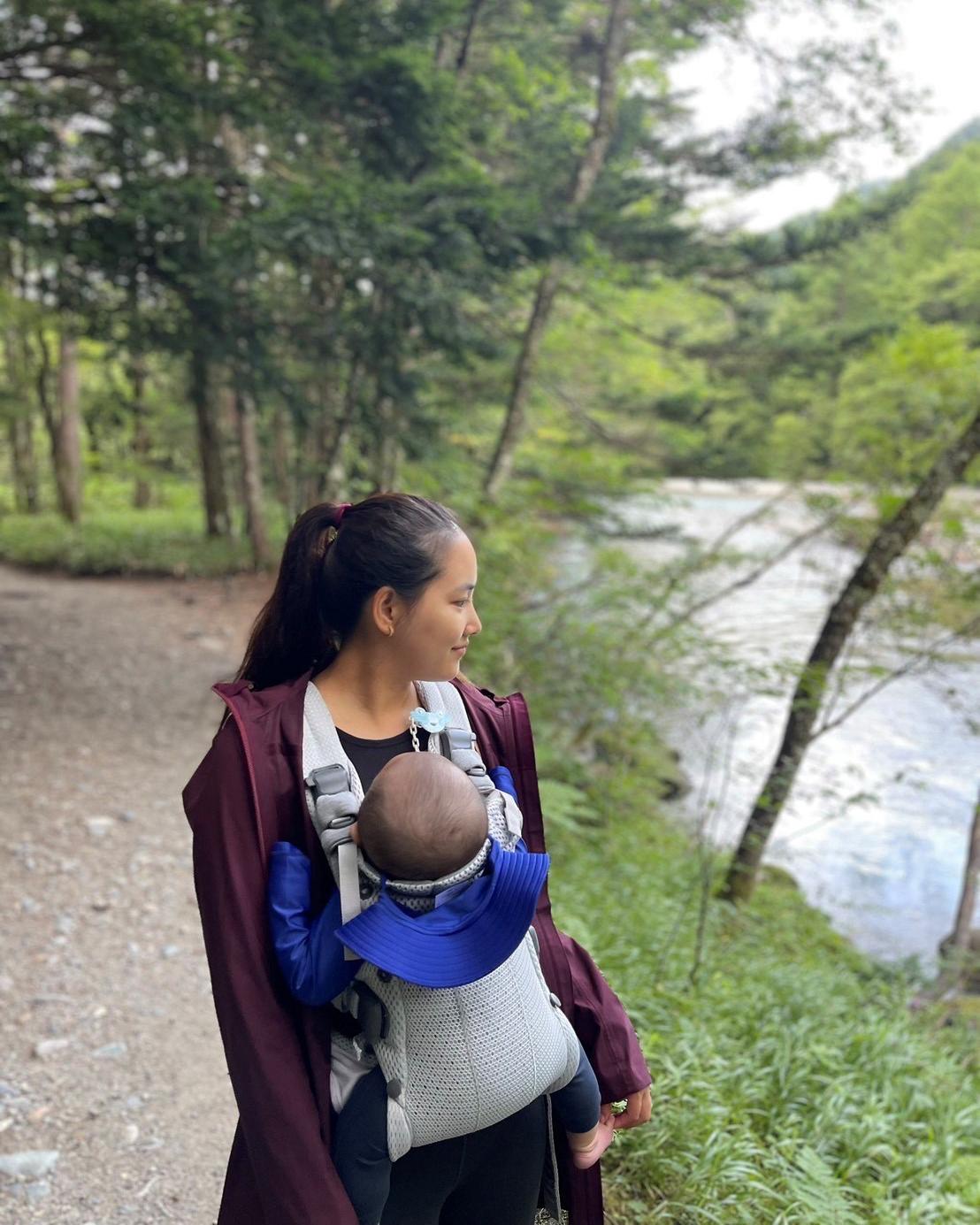 A woman stands on a forest path, carrying a baby in a front carrier, looking at a river. Trees and greenery surround them.