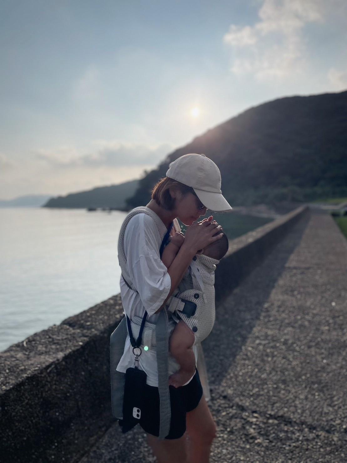 A person wearing a cap holds a baby in a carrier, standing on a path by a body of water with hills in the background under a cloudy sky.