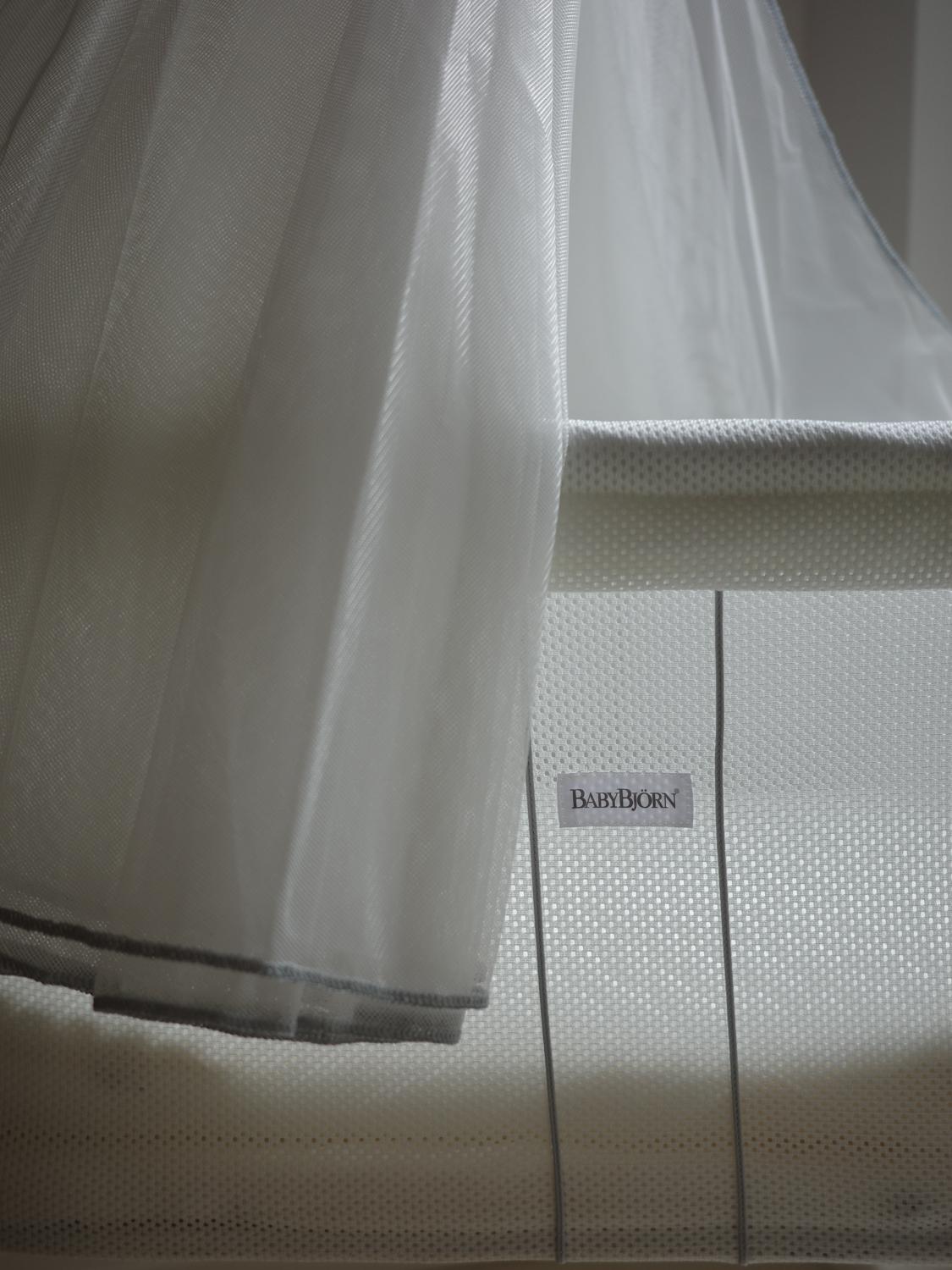 Close-up of a white BabyBjörn bassinet with a sheer canopy, featuring a mesh fabric and the brand label visible on the side.