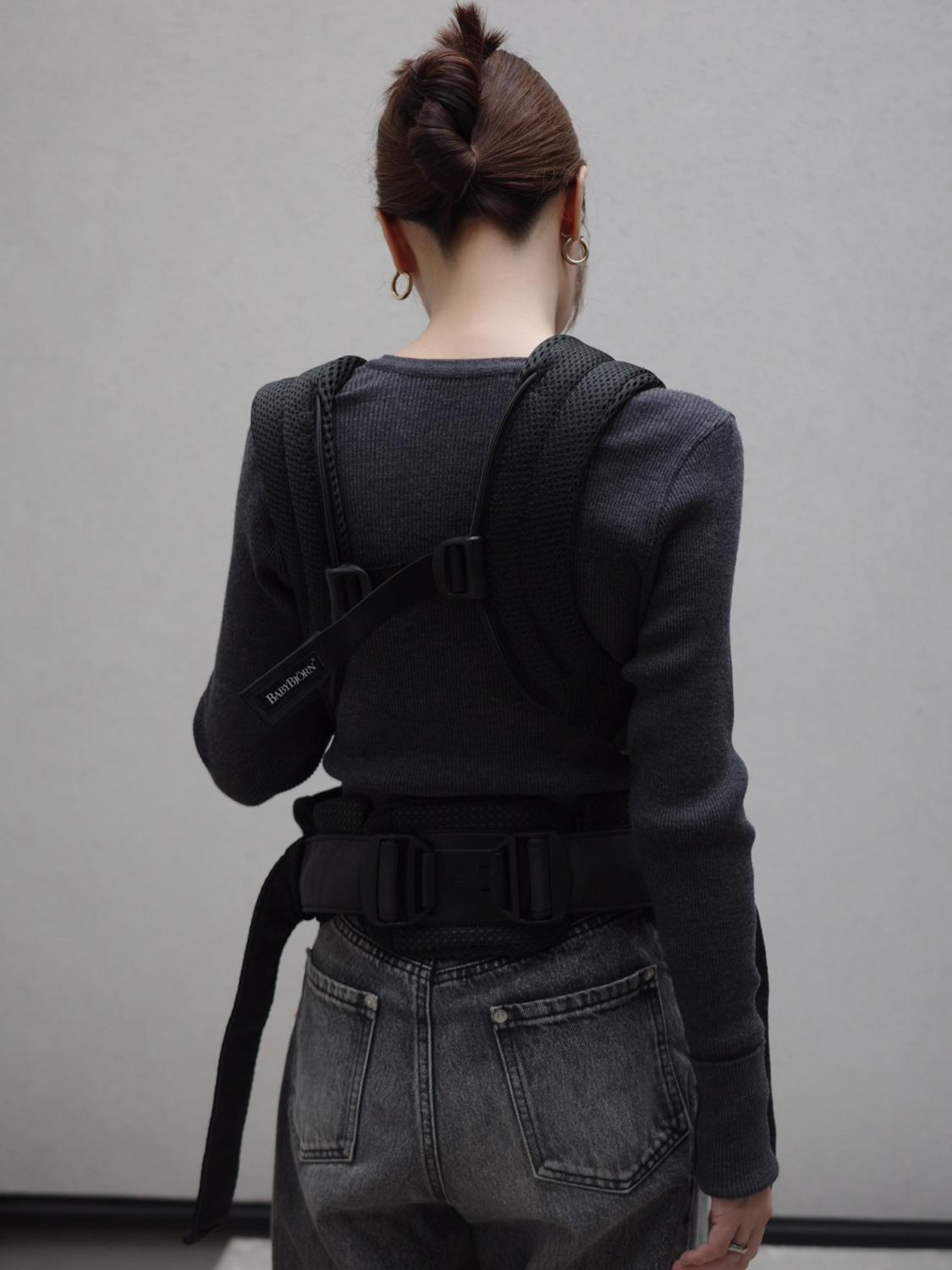 Person with a bun hairstyle wearing a dark sweater and jeans, seen from the back, with a black harness strapped over their shoulders.