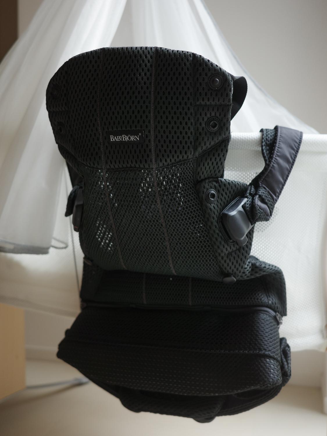 Black mesh baby carrier with adjustable straps, hanging in front of a white curtain.