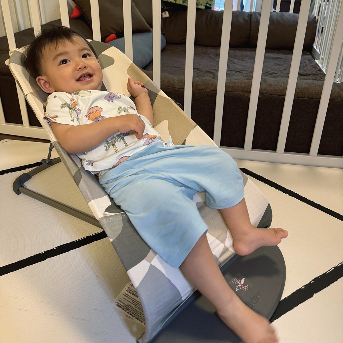 A smiling toddler in a white shirt and blue pants reclines in a baby bouncer, surrounded by a white playpen.