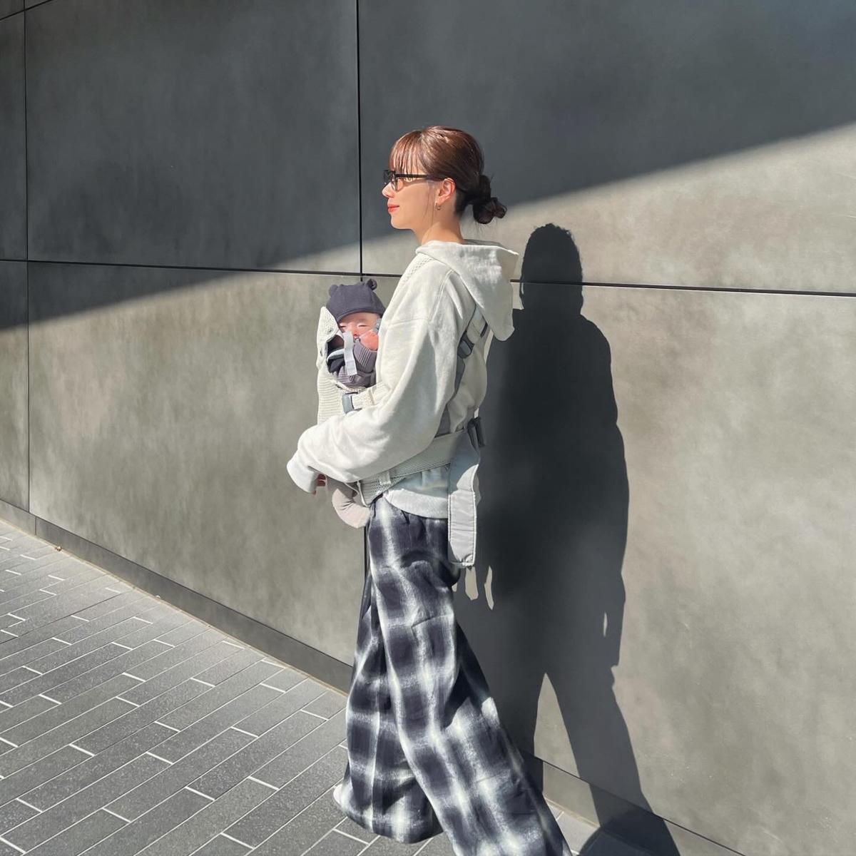 Woman with a bun and sunglasses carries a baby in a front carrier, walking along a sunlit, modern concrete wall.