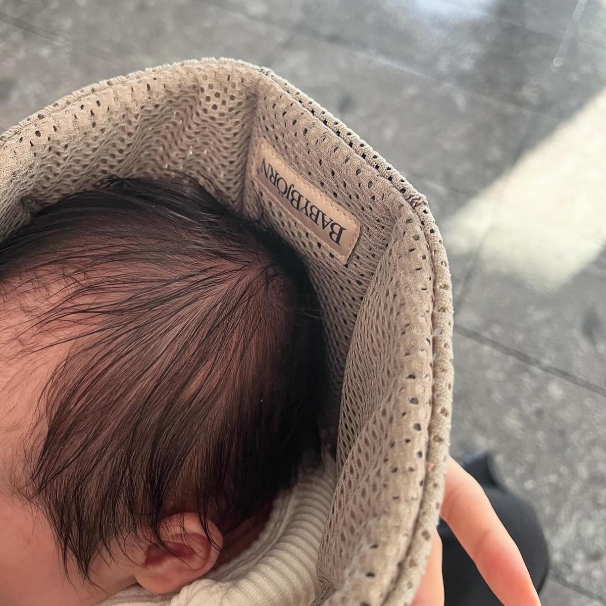 Close-up of a baby resting in a beige, textured carrier. The label reads "BABYBJÖRN." The background is a dark, shiny surface.