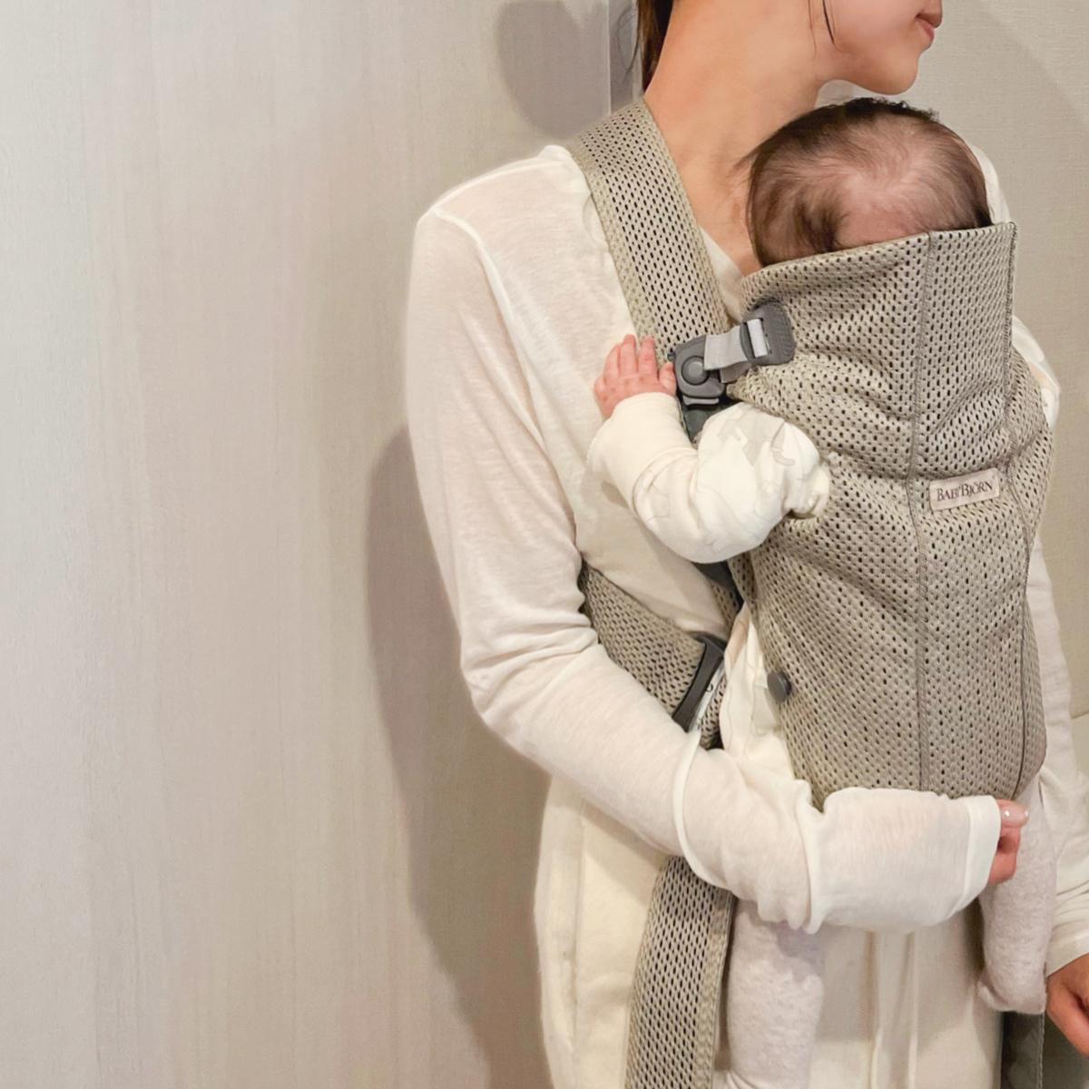 A woman in a white top carries a baby in a beige gray baby carrier, standing against a light-colored wall.