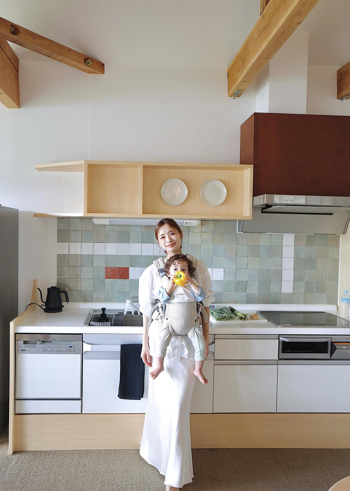 Woman in a white dress holding a baby in a carrier, standing in a modern kitchen with wooden accents and tiled backsplash.