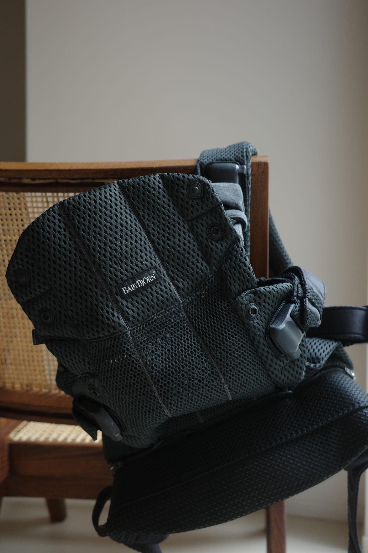 Black mesh baby carrier draped over a wooden chair with a woven backrest, in a softly lit room.