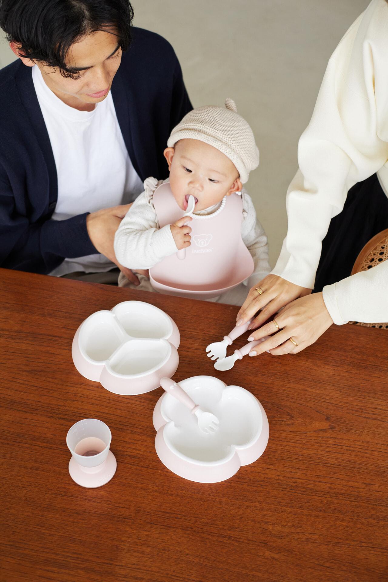 Baby in a bib sits between two adults at a table with pink baby dishes, holding a spoon.
