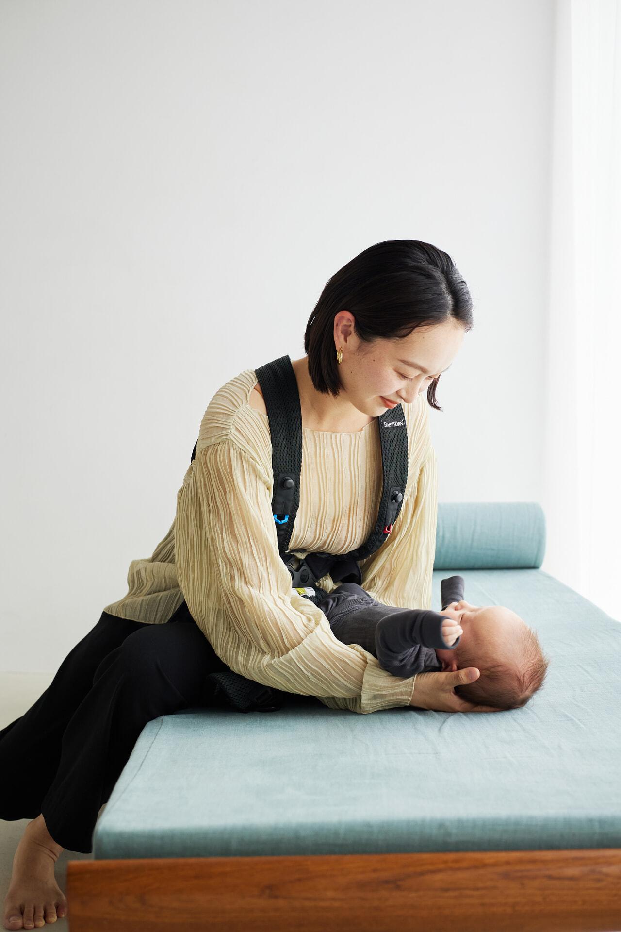 Woman sitting on a bench, smiling at a baby lying on her lap. She wears a beige top and black pants, and the baby is in dark clothing.