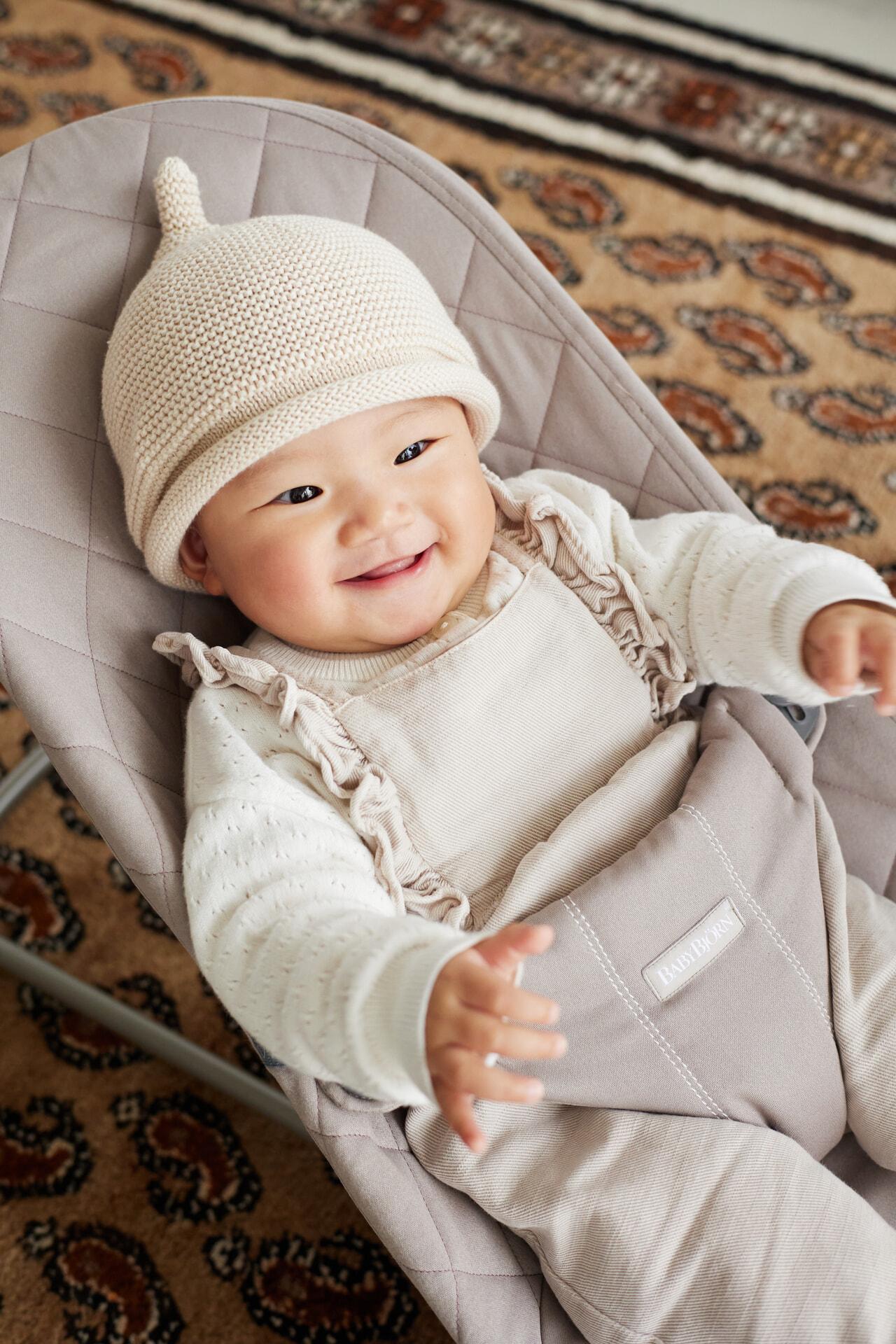 A smiling baby wearing a knitted hat and cozy outfit sits in a cushioned seat on a patterned rug.