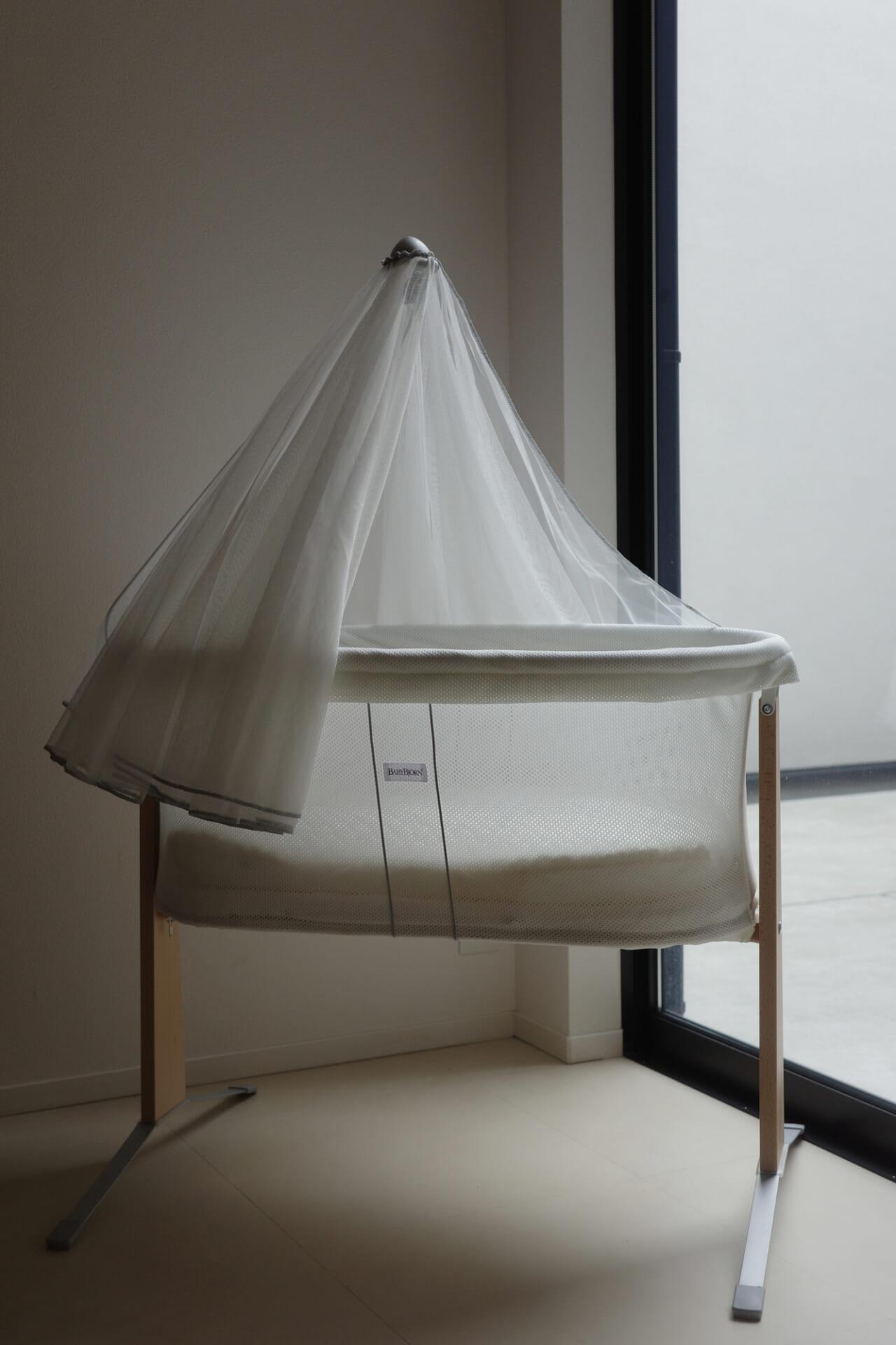A minimalist baby bassinet with a sheer canopy stands by a large window, allowing natural light to illuminate its modern design.