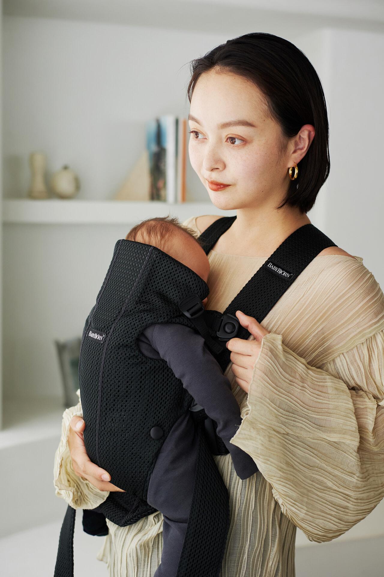A woman holds a baby in a black carrier. She wears a beige dress and stands in a softly lit room with shelves in the background.