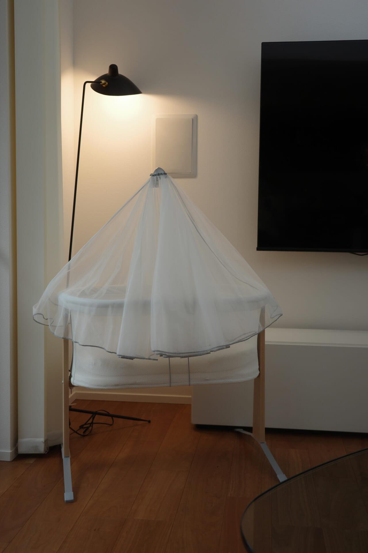 A white bassinet with a sheer canopy is placed near a floor lamp and a wall-mounted TV on a wooden floor.