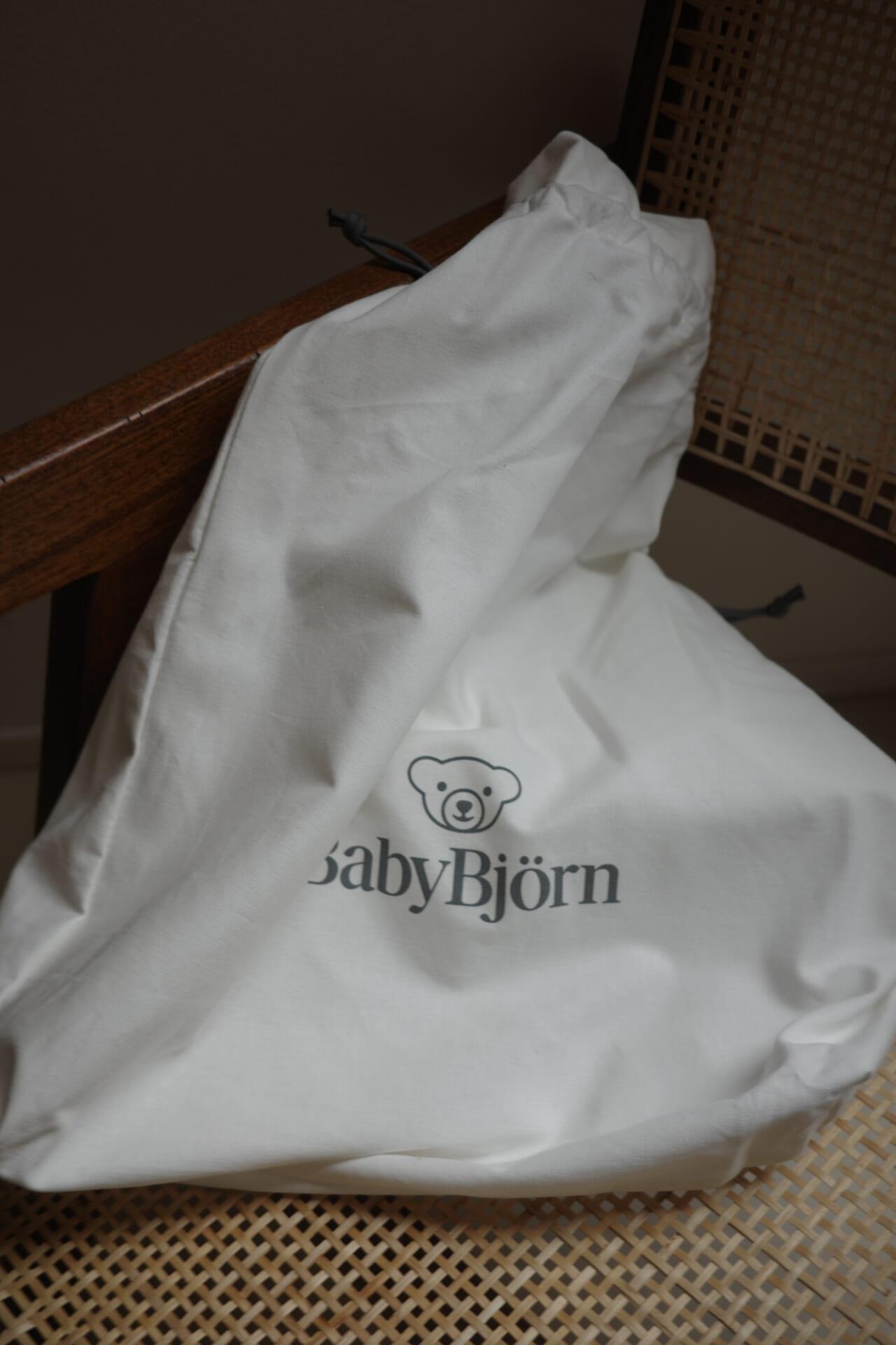 White BabyBjörn bag with a bear logo, resting on a wooden and woven chair.