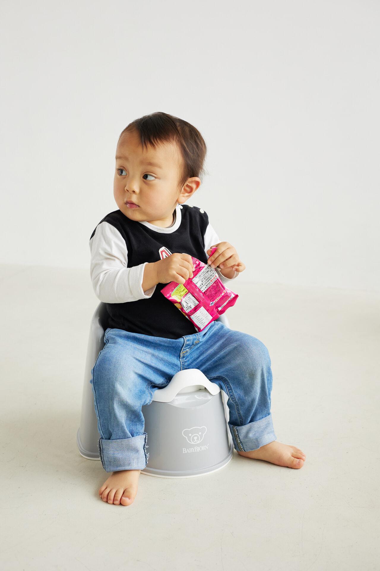 A toddler sits on a gray potty, holding a colorful snack package, wearing a black and white shirt with blue jeans, looking to the side.