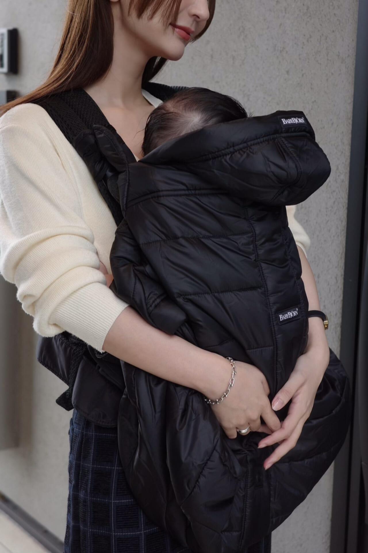 Woman holding a baby in a black carrier with a winter cover wearing a cream sweater and dark skirt, standing against a gray wall.