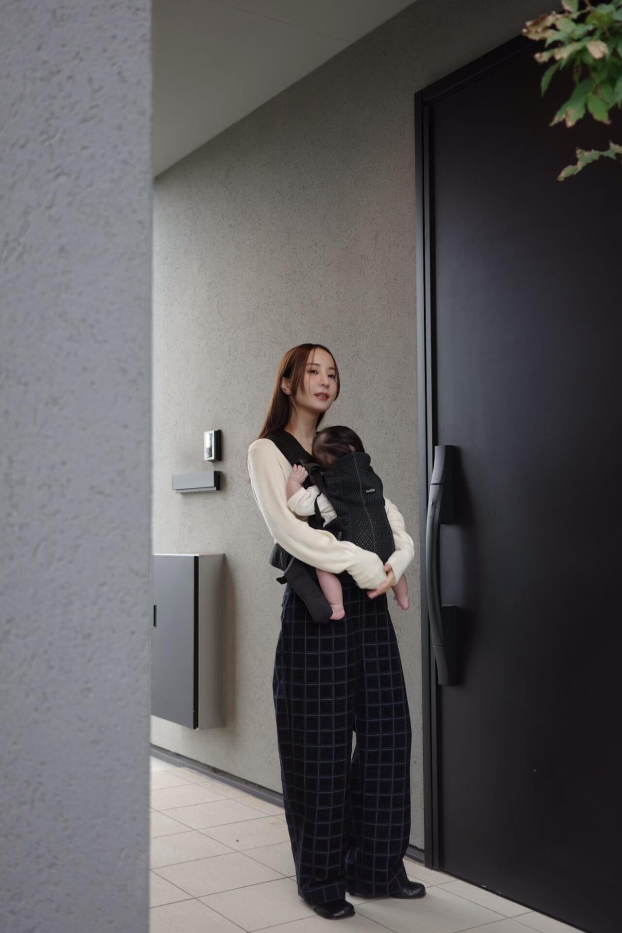 Woman standing by a modern door, wearing a cream sweater and checkered pants, carrying a baby in a black carrier.