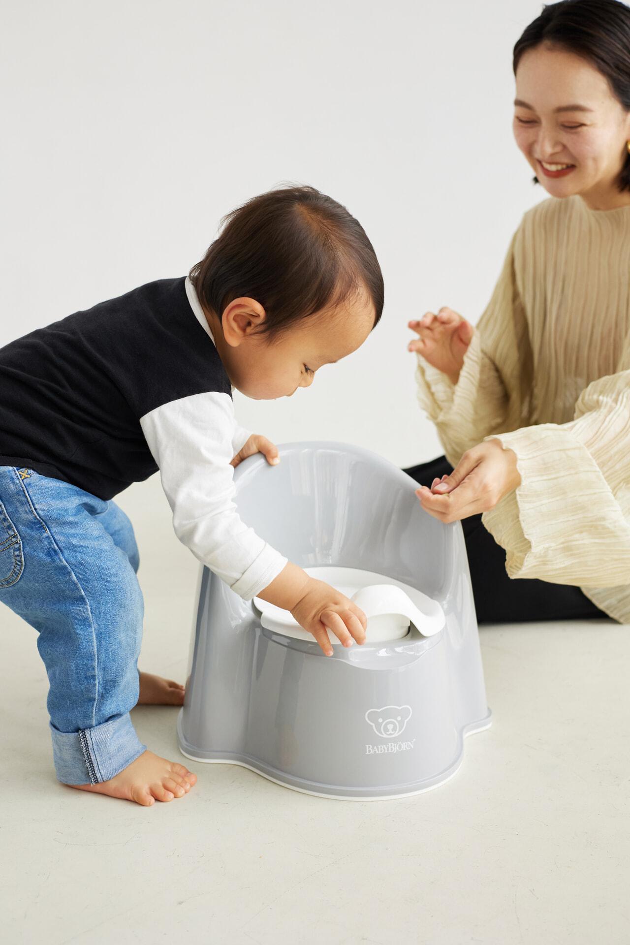 A toddler interacts with a gray potty chair while a smiling adult watches, sitting on the floor.