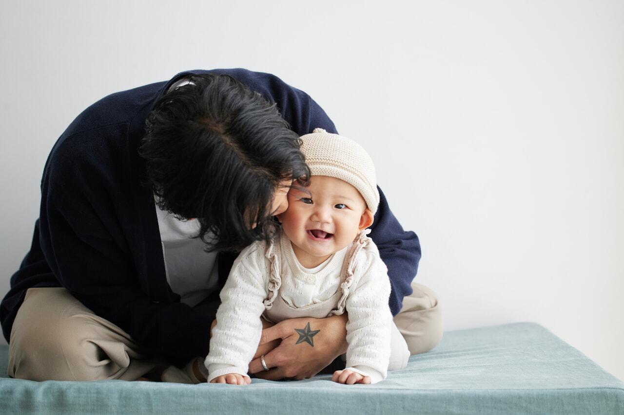 Adult embracing a smiling baby wearing a knit hat, both sitting on a light blue surface against a plain background.