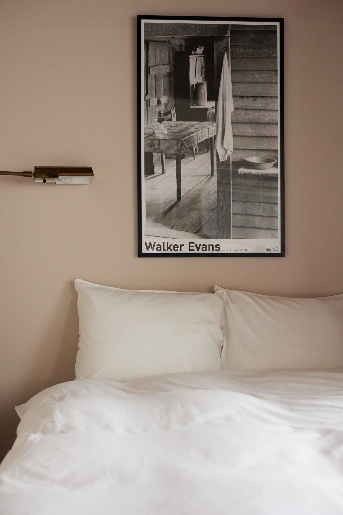 A neatly made bed with white pillows and sheets is beneath a wall-mounted black-and-white Walker Evans photograph in a simple, modern bedroom.