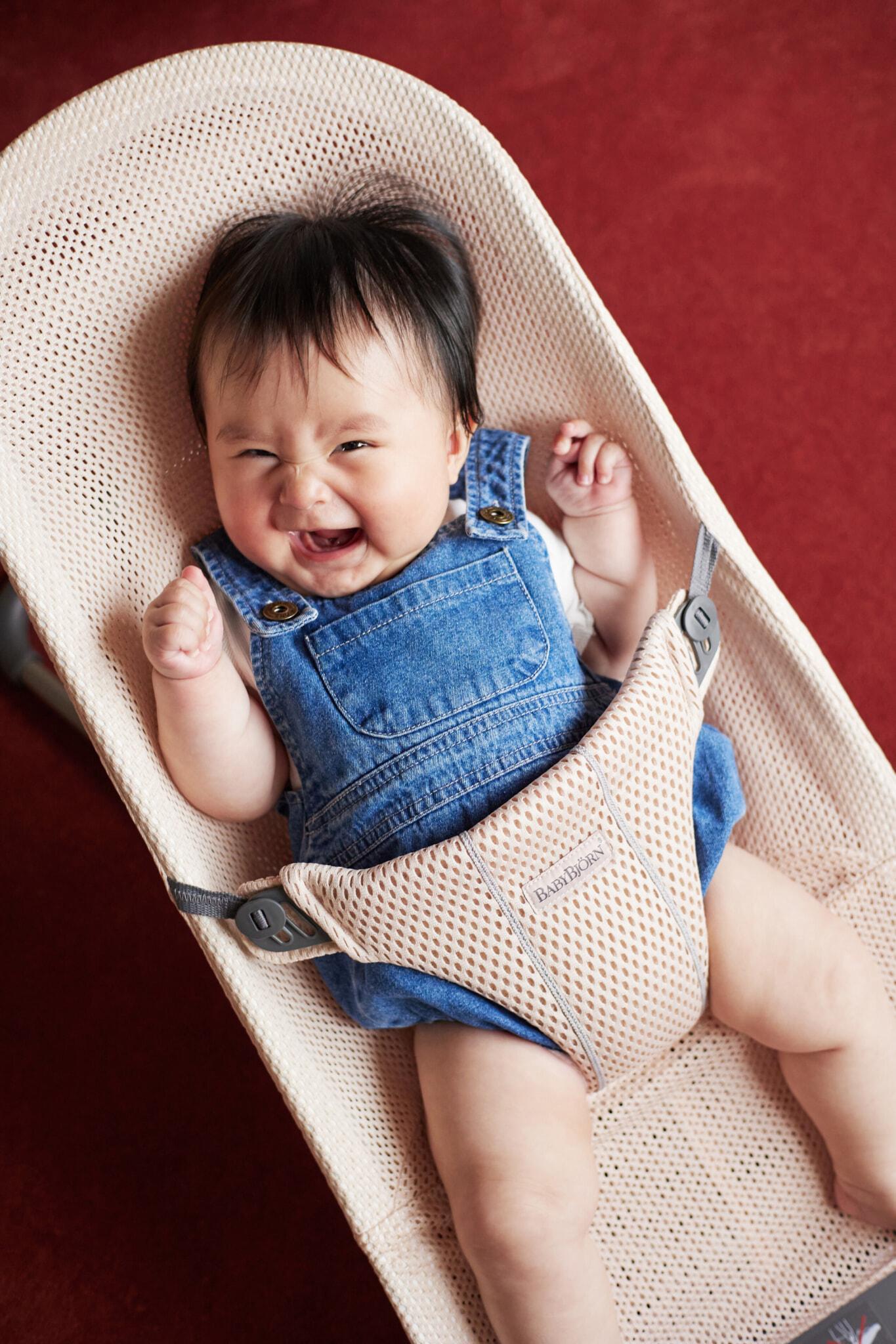 A smiling baby wearing denim overalls sits in a beige baby bouncer on a red carpet.