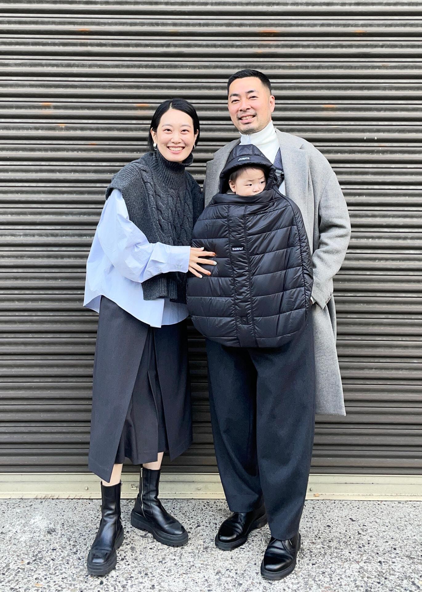 A smiling couple stands with a child wrapped in a black coat. They're in front of a corrugated metal wall, dressed in stylish neutral outfits.