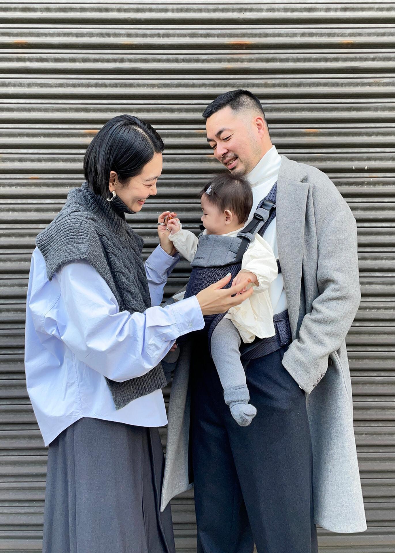 A couple smiles at their baby, who is in a carrier worn by the father. They stand in front of a corrugated metal wall.