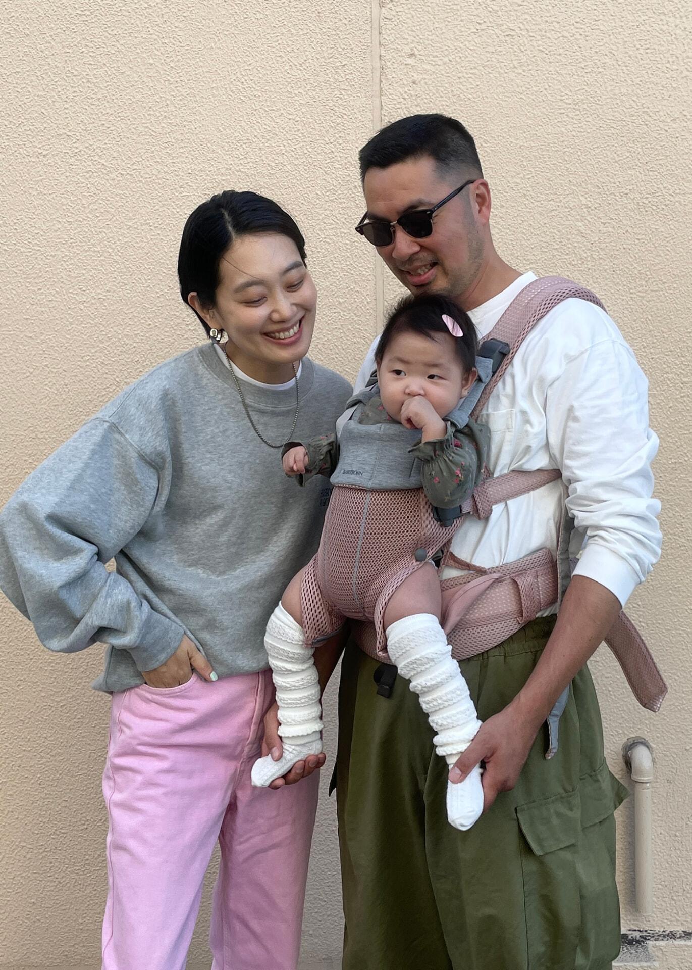 A smiling couple stands with a baby in a carrier. The woman wears a gray sweater and pink pants; the man wears sunglasses and a white shirt.