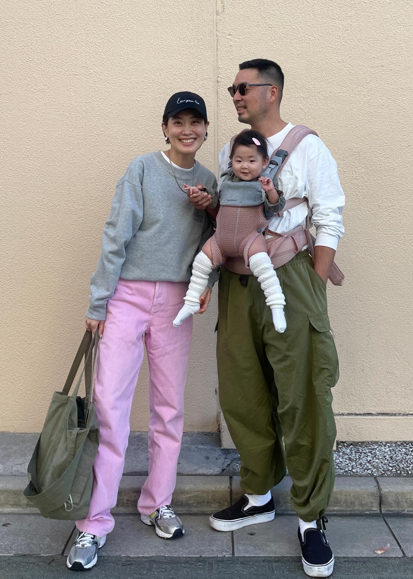 A smiling couple poses with their baby in a carrier. The woman wears pink pants and a cap, while the man wears green pants and sunglasses.