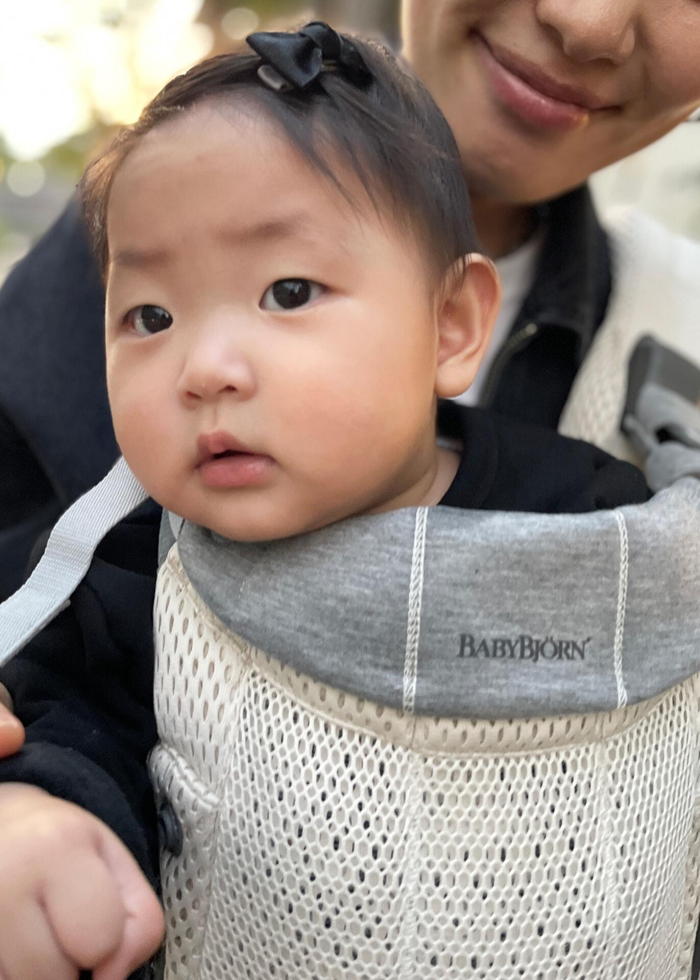 A baby in a BabyBjörn carrier, wearing a black outfit and a small black bow, held by an adult.