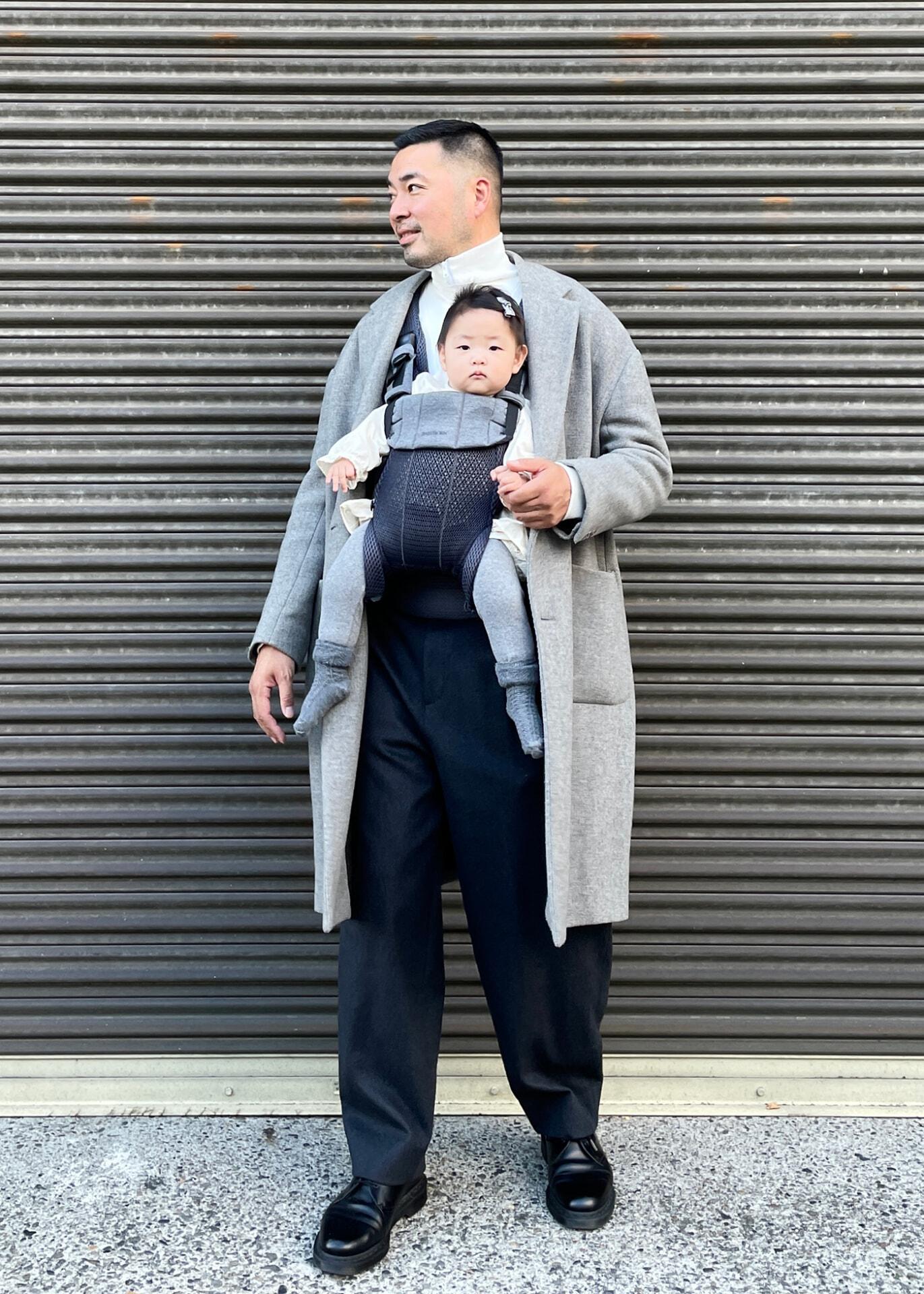 A man in a gray coat stands in front of a corrugated metal background, carrying a baby in a gray outfit and carrier.