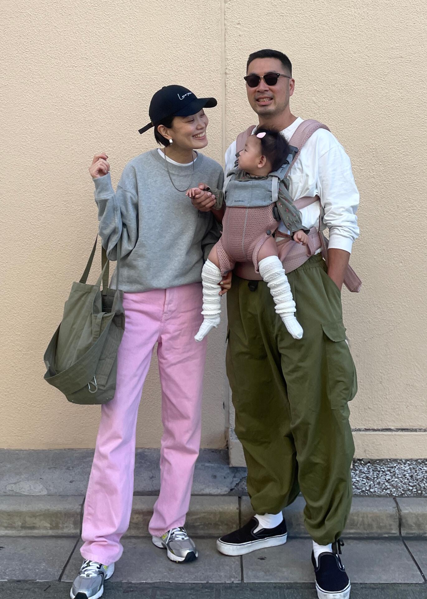 A smiling couple stands with a baby. The woman wears a cap, gray sweater, and pink pants. The man wears sunglasses and carries the baby in a front carrier.
