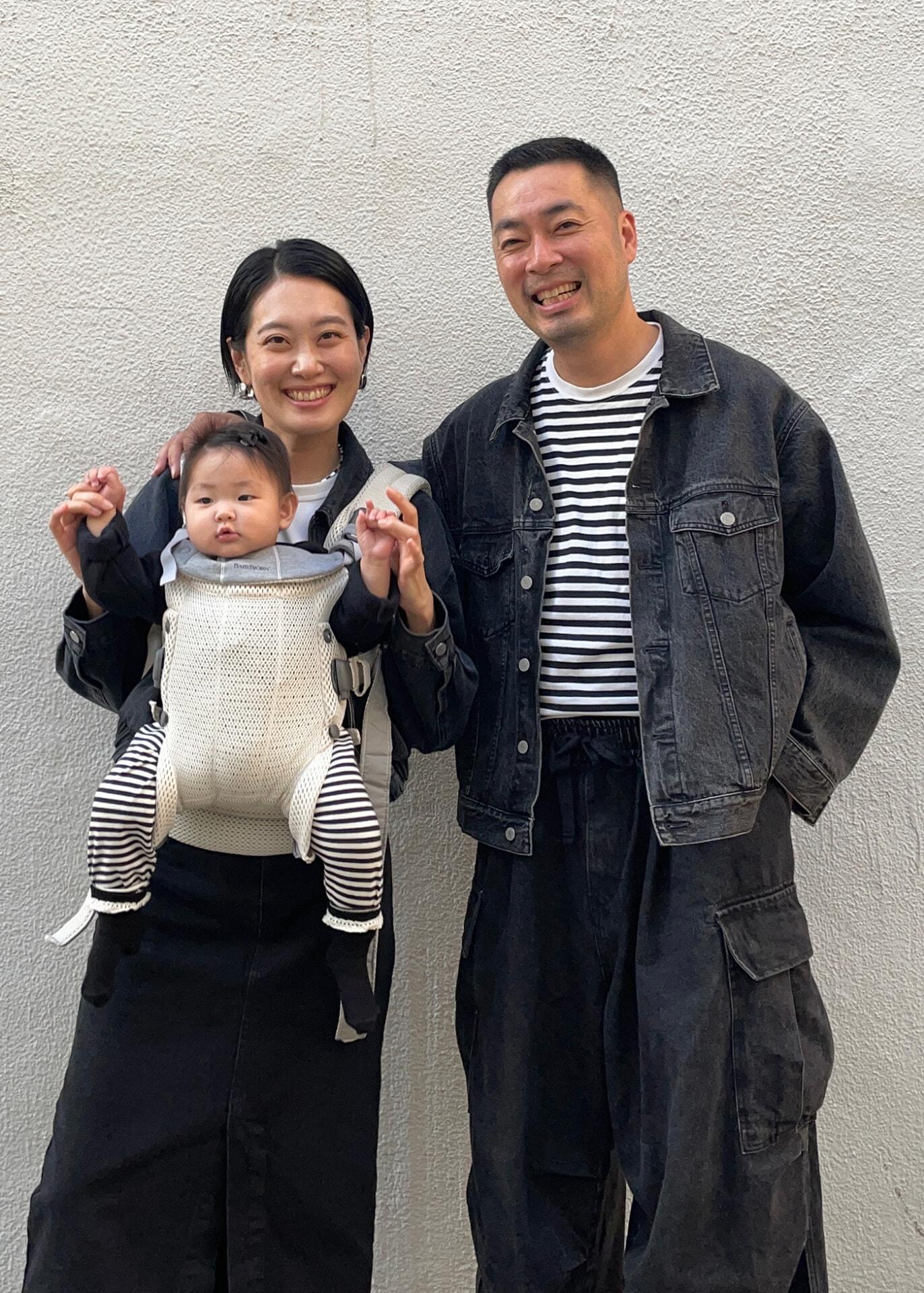 A smiling couple stands against a gray wall, holding a baby in a front carrier. All wear black and white outfits.