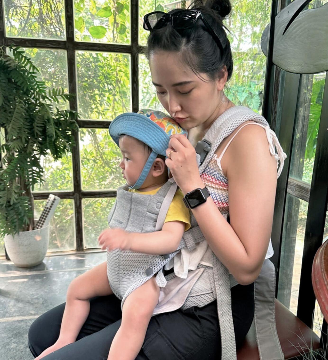 A woman sits indoors, holding a baby in a carrier. The baby wears a blue hat. Sunlight filters through large windows with greenery outside.