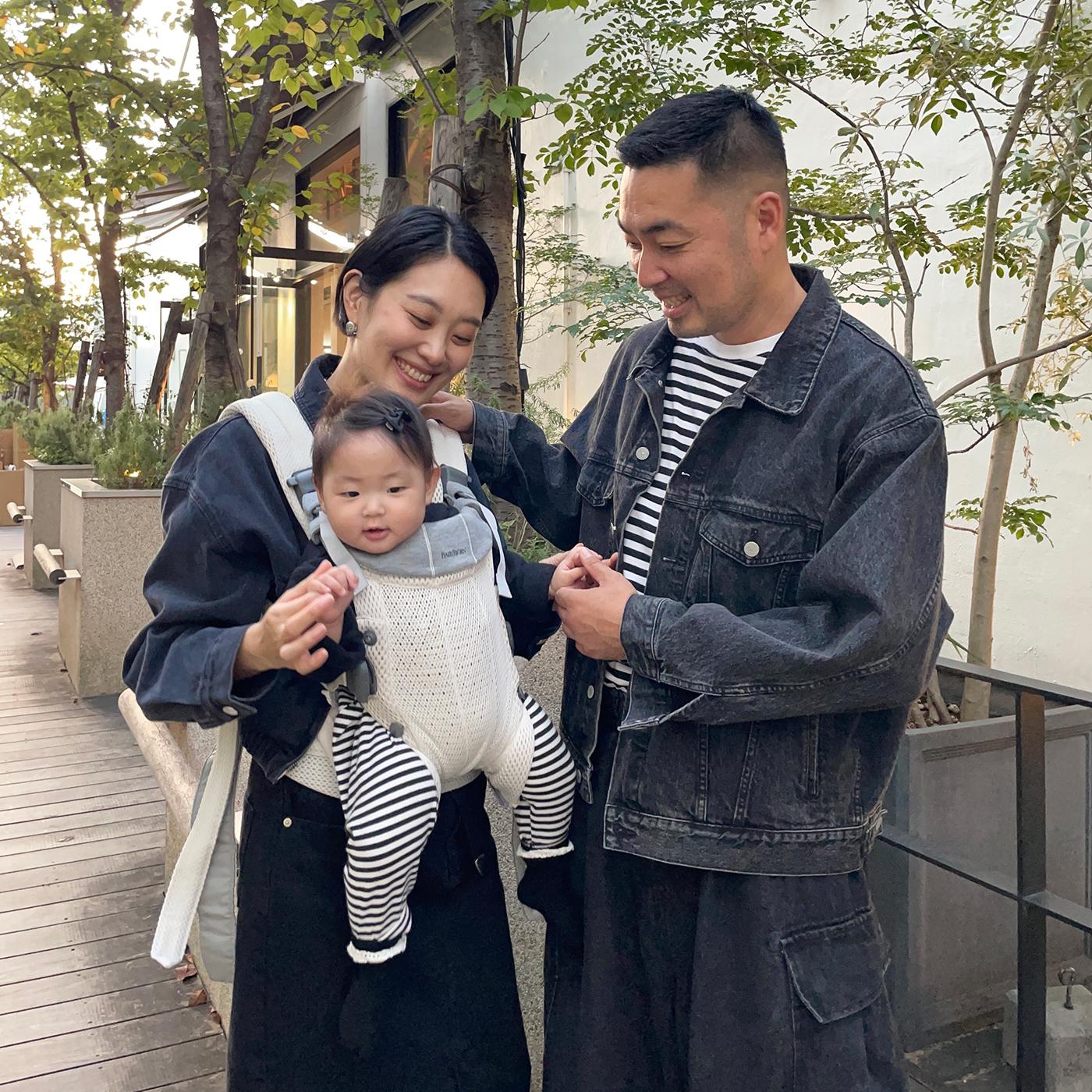 A smiling couple stands on a wooden path with their baby in a carrier. They're wearing matching dark denim outfits, surrounded by greenery.