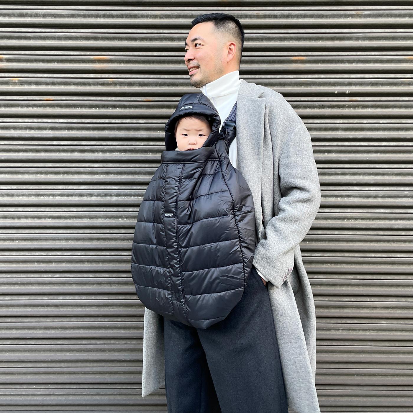 Man in a gray coat carries a baby in a black padded carrier, standing against a metal shutter background.