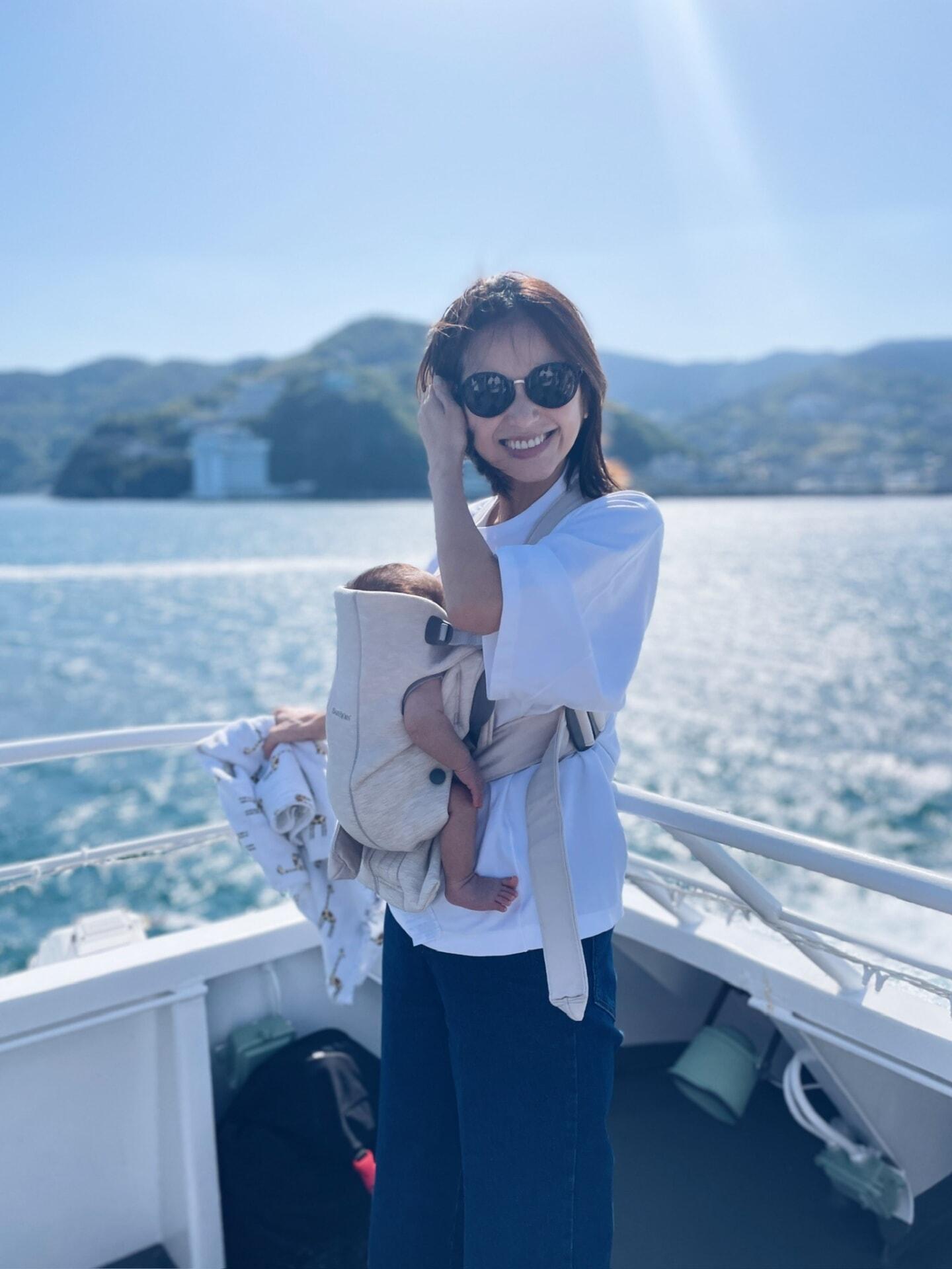 A woman wearing sunglasses holds a baby in a carrier on a boat, with scenic mountains and water in the background.