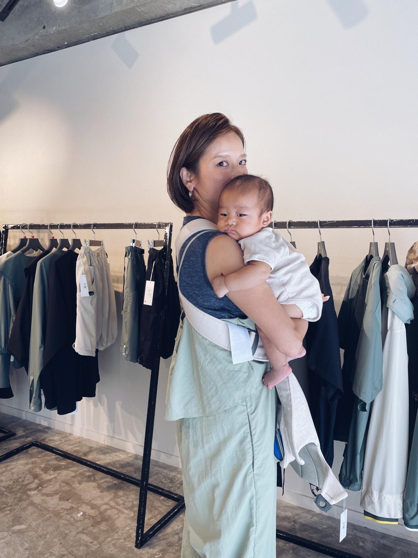 A woman carrying a baby in a store, surrounded by hanging clothes on racks. The baby is resting on her shoulder.