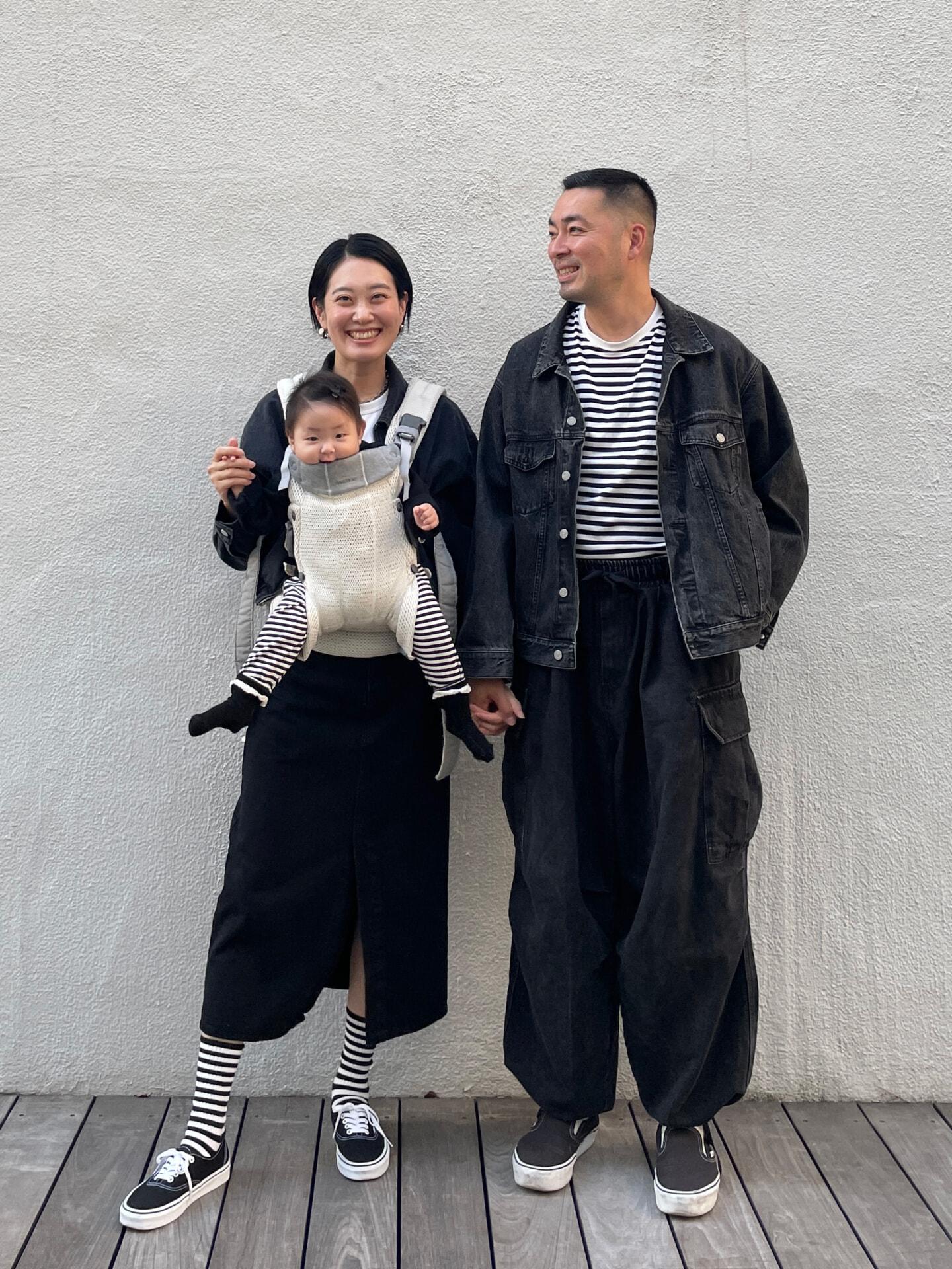 A smiling couple stands with a baby in a carrier. They all wear matching black and white striped outfits and black shoes against a plain wall.