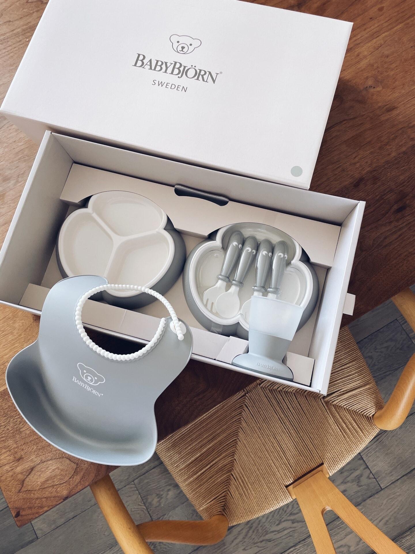 BabyBjörn feeding set in a box on a wooden table, including a bib, plates, utensils, and a cup, all in matching gray tones.