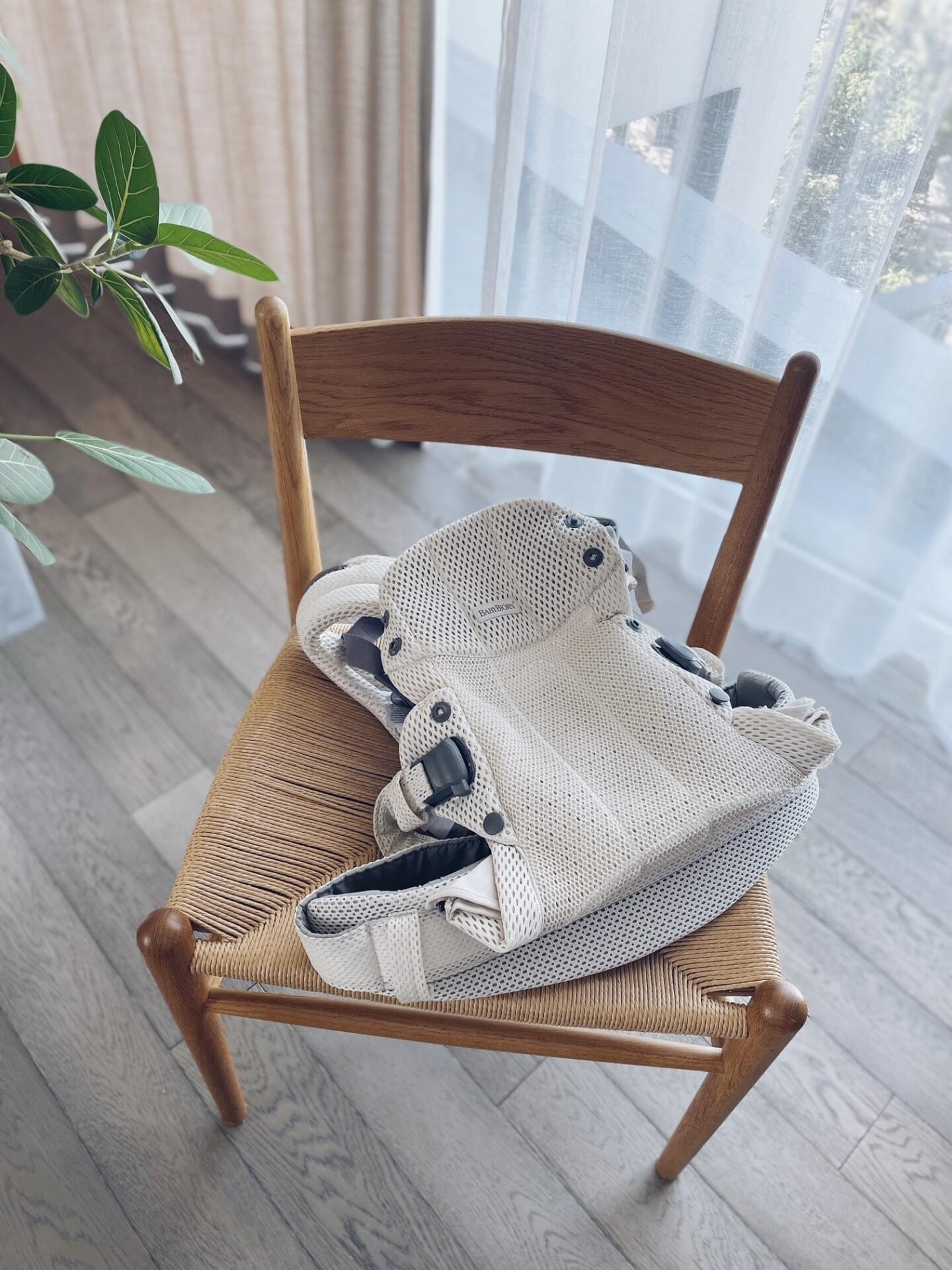 A beige baby carrier with mesh fabric is placed on a wooden chair with woven seat, near a window with sheer curtains and a potted plant.